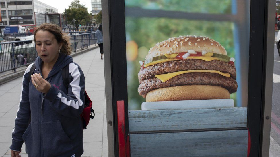 Woman eating a burger while passing an advertisement for burgers at Elephant and Castle in London, UK