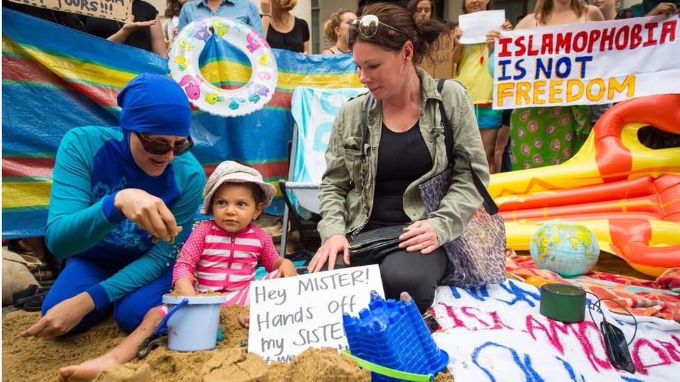 Two women, one wearing a burkini and one not, sit next to a child playing in the sand; a sign reads 'hey mister hands off my sister'