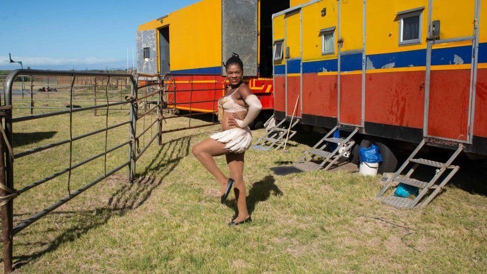 A dancer posing in Cape Town. She is outside standing in what looks like a field in front of a colourful cable home.