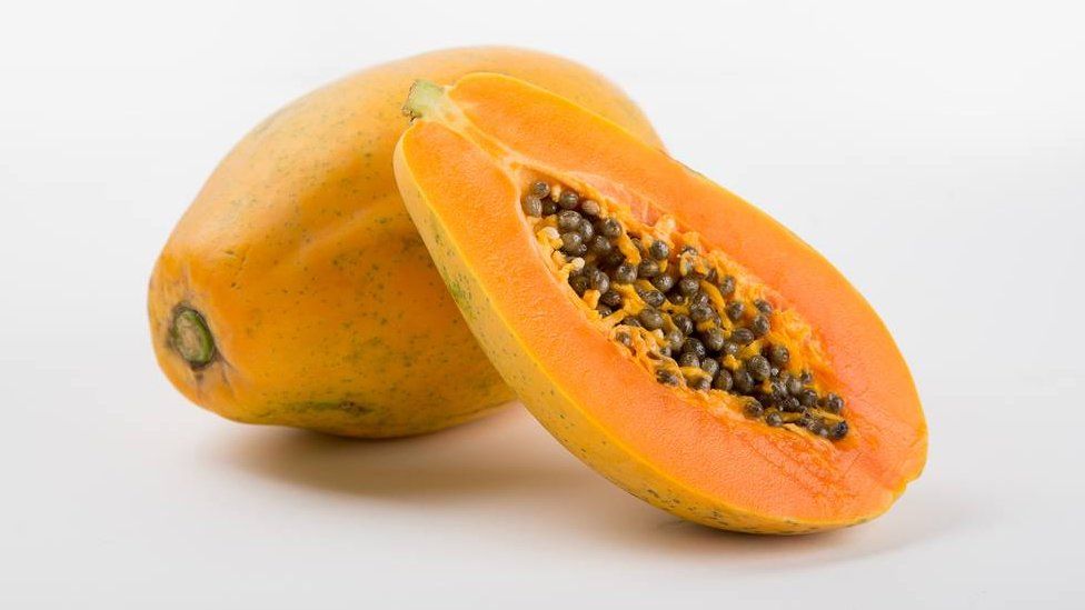 A photo of the maradol papaya - a large oval fruit with an orange colour. One fruit is cut in half, showing the dark seeds clustered in its core