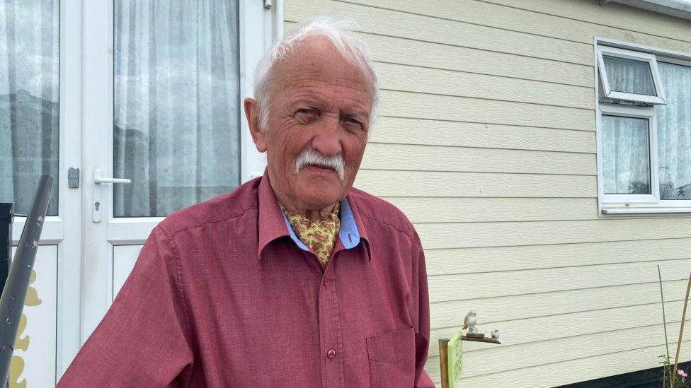 Tom Bird stand in front of his rental home. He is wearing a dark pink shirt and a yellow cravat. He has white hair and a white moustache.