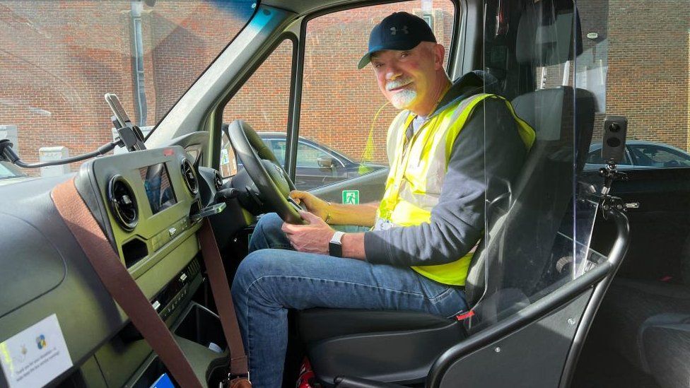 A man with a short grey beard sits in the driver's seat of a minibus wearing a hi-vis jacket