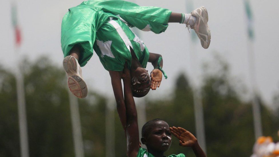 Two acrobatic boys performing in Abuja. They are wearing green and white outfits - the colours of the Nigerian flag - with stars on them. one boy is holding another with just one hand above his head. They are both saluting. They have white face paint on and a serious expression.