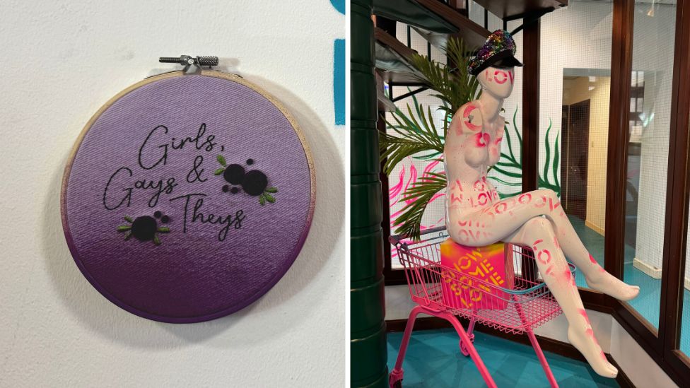 Cross stitch sign reading 'Girls gays and theys' and a spray painted mannequin sat on a hot pink trolley