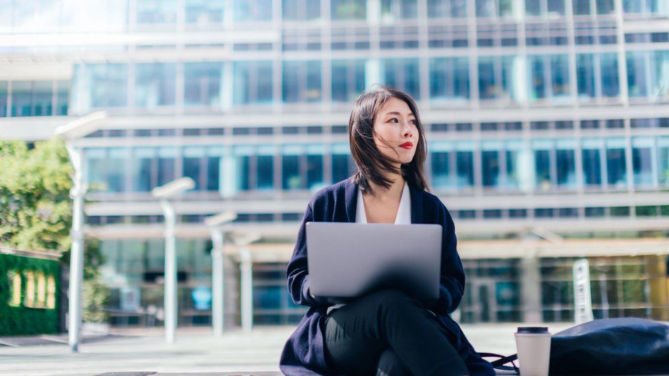 Female worker on laptop near corporate buildings.