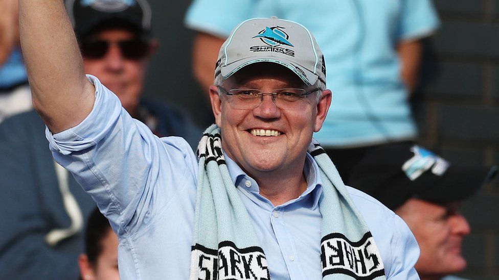 Scott Morrison wearing a baseball cap and rugby scarf waves to the crowd while celebrating his victory at a rugby league match in Sydney on Sunday