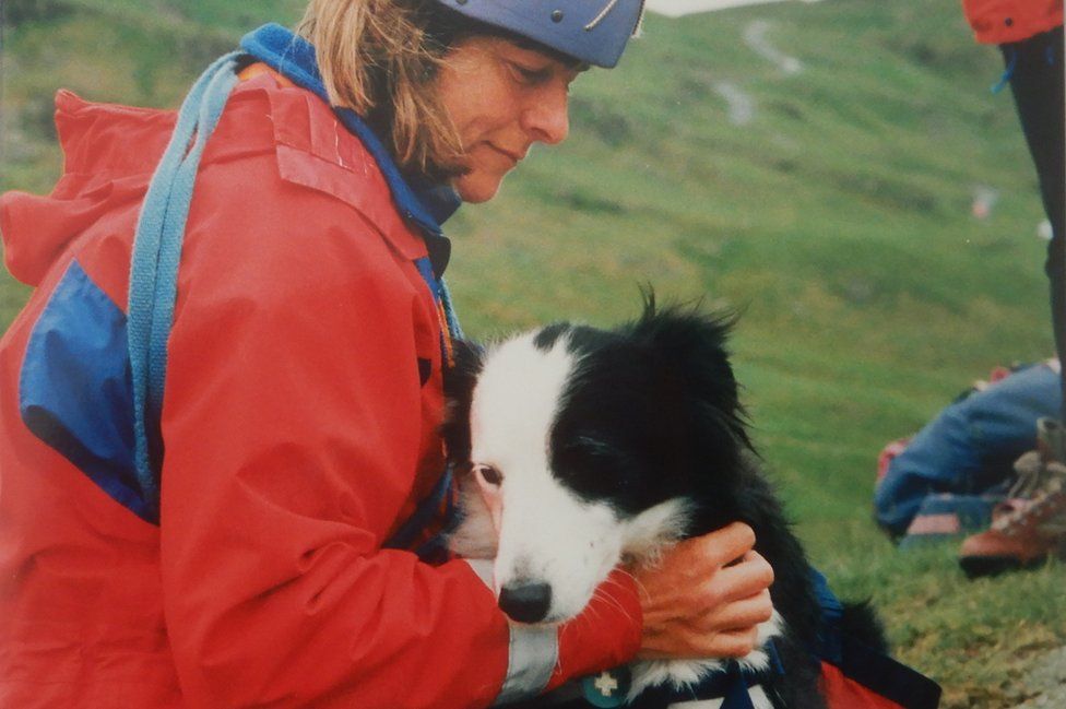 A woman in red coat and climbing hat holds a collie