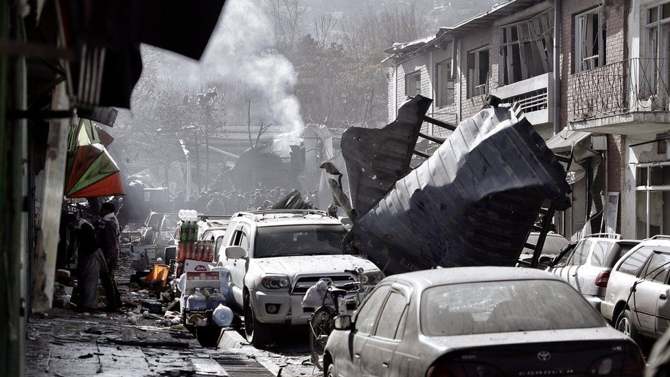 mangle structures blown halfway down a street, with debris all over the pavements and people crowding around a plume of smoke in the background