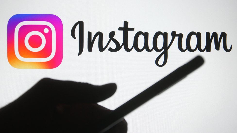 The silhouette of a hand holding a smartphone is seen in front of the Instagram logo