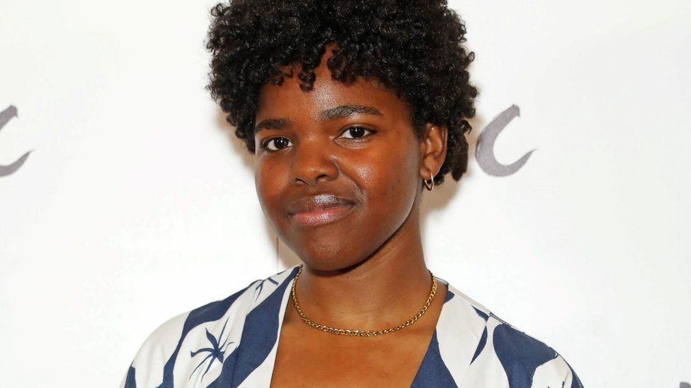 Francesca Amewudah-Rivers. Francesca is a black woman in her 20s with short afro hair and brown eyes. She smiles at the camera, wearing a blue and white blouse and small gold hooped earrings and gold chain necklace.