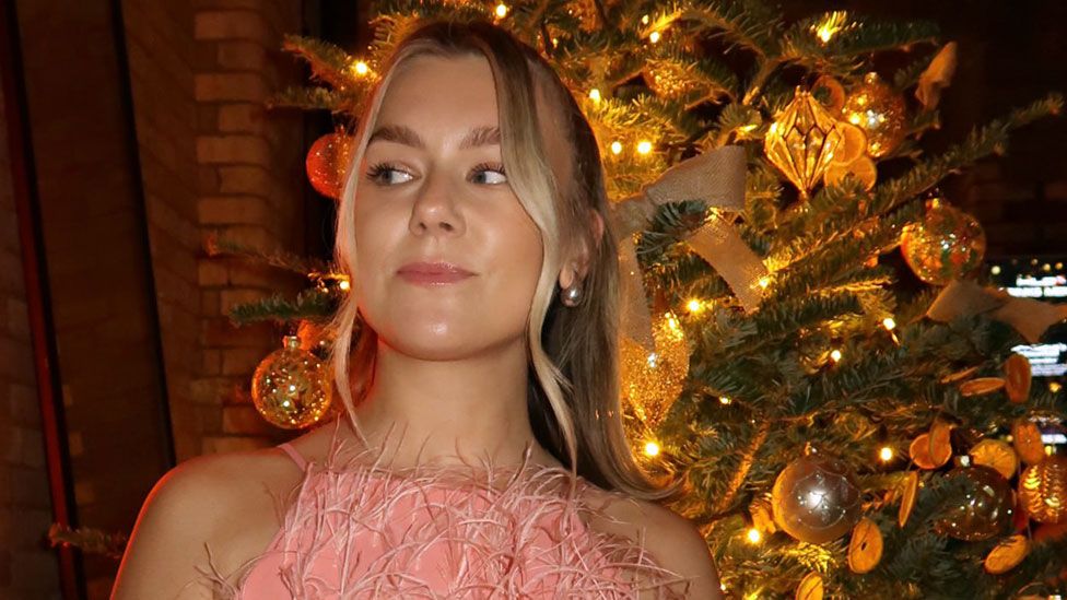 Kali Thompson at a Christmas Party. Kali is a 25-year-old white woman with blonde hair which she wears tied back in a pony tail. she wears a feathered pink dress and is pictured in front of a Christmas tree