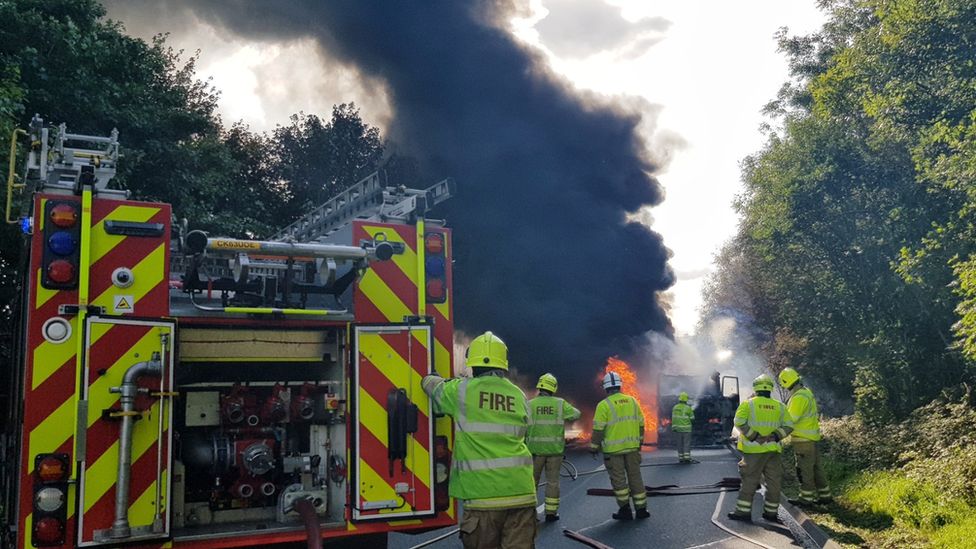 Fire engine with lots of black smoke and flames surrounding a burnt out lorry
