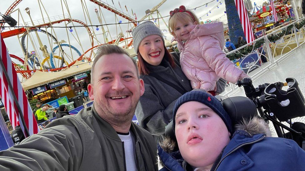 A family of four including a man, woman, a two year old girl and a ten year old boy in a wheelchair, smile for a selfie in front of an amusement park