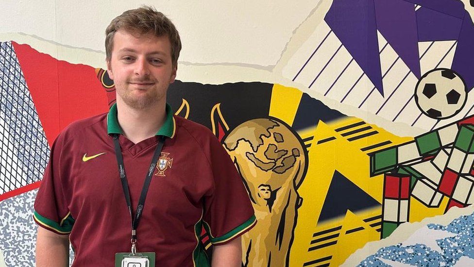Josh Phillips is wearing a Portuguese football shirt, it's maroon with a yellow and green striped collar. He's standing in front of a very bright, multi coloured background which has a picture of the world cup and a football.