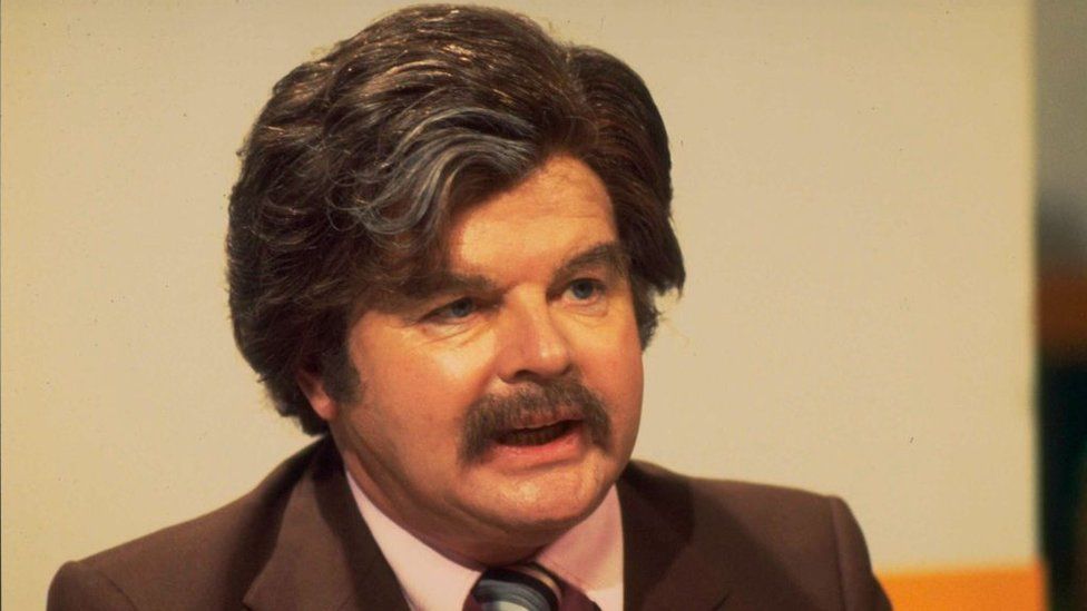 El cómico Benny Hill vestido como Dickie Davies en 1979, con un peinado similar y un bigote poblado
