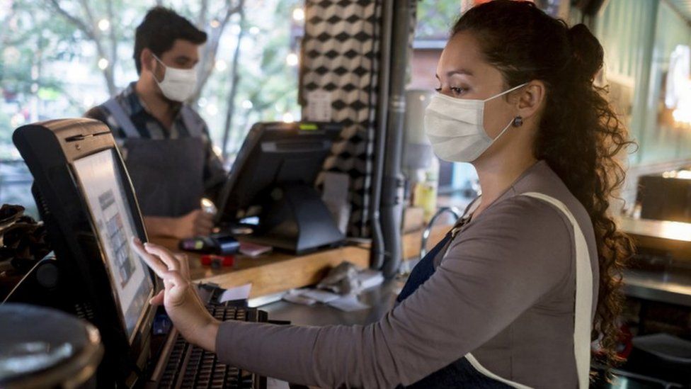 A woman working at the cashier at a restaurant wearing a facemask
