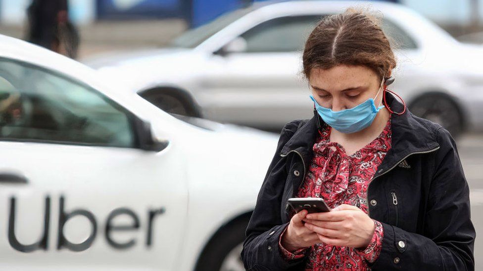 A woman wearing a face mask uses a phone as an Uber passes by in the background.