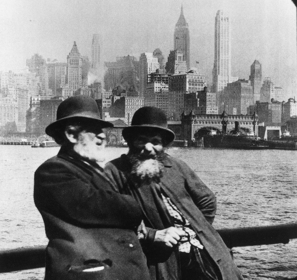 Two Jewish men lean against a barrier with the New York skyline behind them