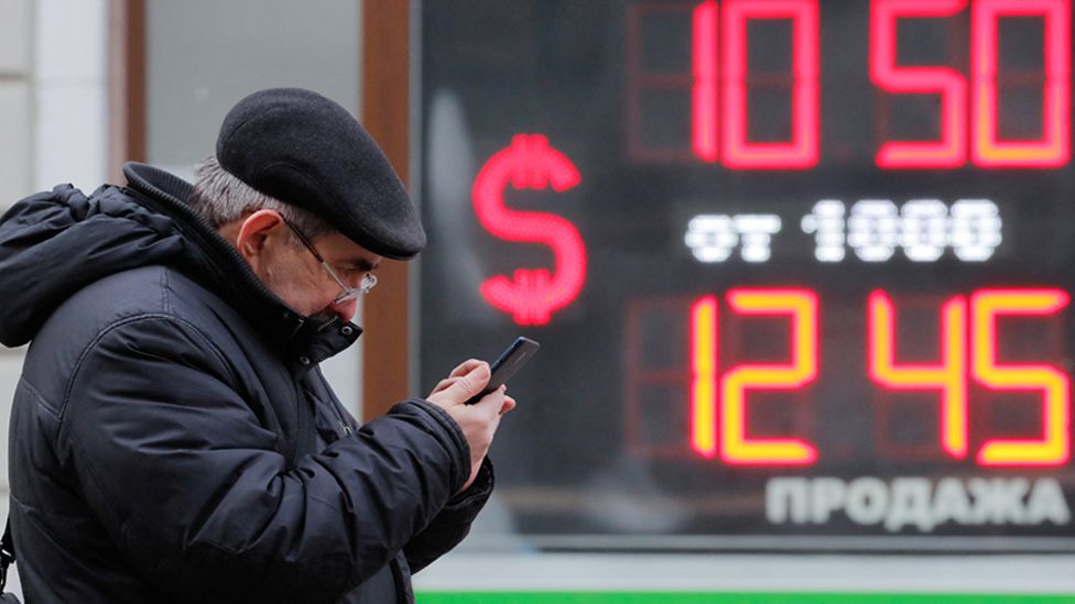 A man uses his smartphone near a board showing currency exchange rates of US dollar against the Russian rouble in Saint Petersburg, Russia 28 February 2022