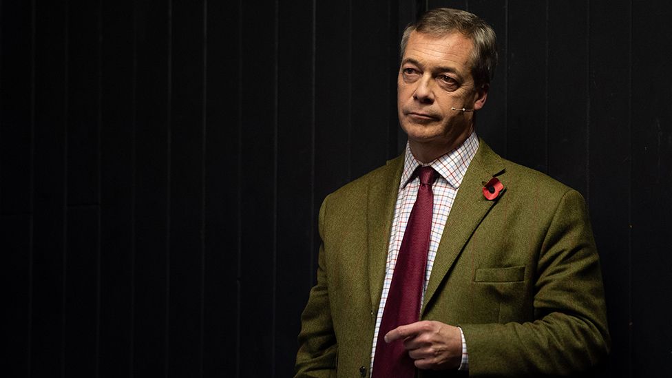 Nigel Farage wears a dark green suit with a white shirt, patterned with criss-crossing red and white lines, underneath. He wears a burgundy neck tie and has a paper poppy pinned to his left lapel. He appears to be on a stage, he's illuminated by a light but the space around him is dark. A microphone/earpiece is just visible, the microphone arm extending from Mr Farage's ear to just above his mouth. He looks as if he's concentrating.