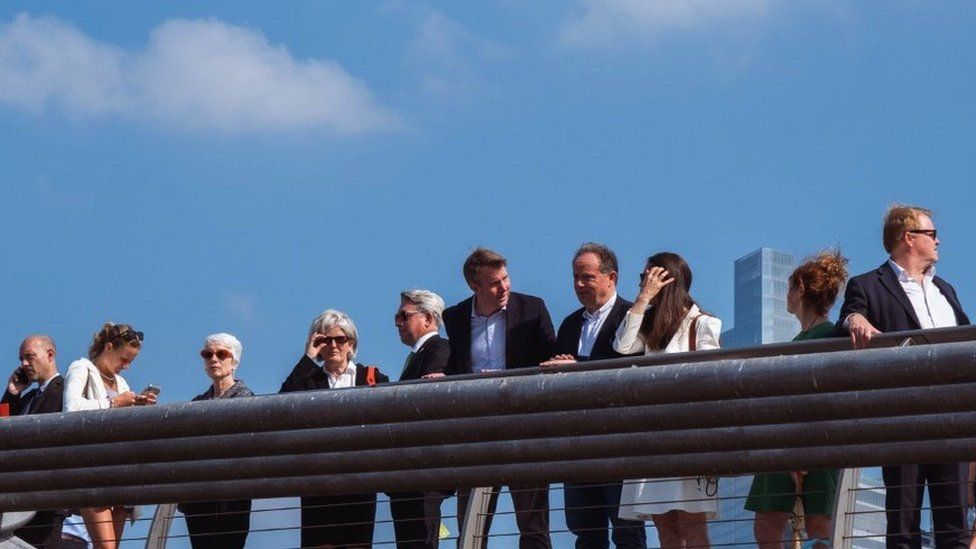 A group of business people lean over a handrail on millennium bridge, bright blue sky in the background and city scape