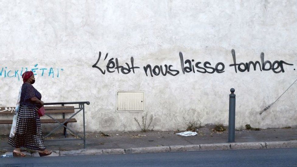 A woman walks in a street in "Les Marronniers" neighbourhood as a handwriting reads 'State lets us down' on the wall, on August 30, 2021
