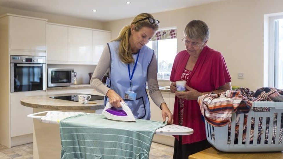 Female carer is ironing in the kitchen to help an elderly woman