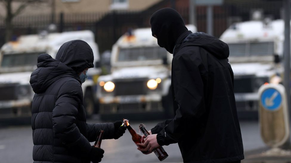 Two young men with their faces covered standing in the foreground, with a row of police Land Rovers in the middle distance. One of them is lighting a petrol bomb which the other one is holding in his hand