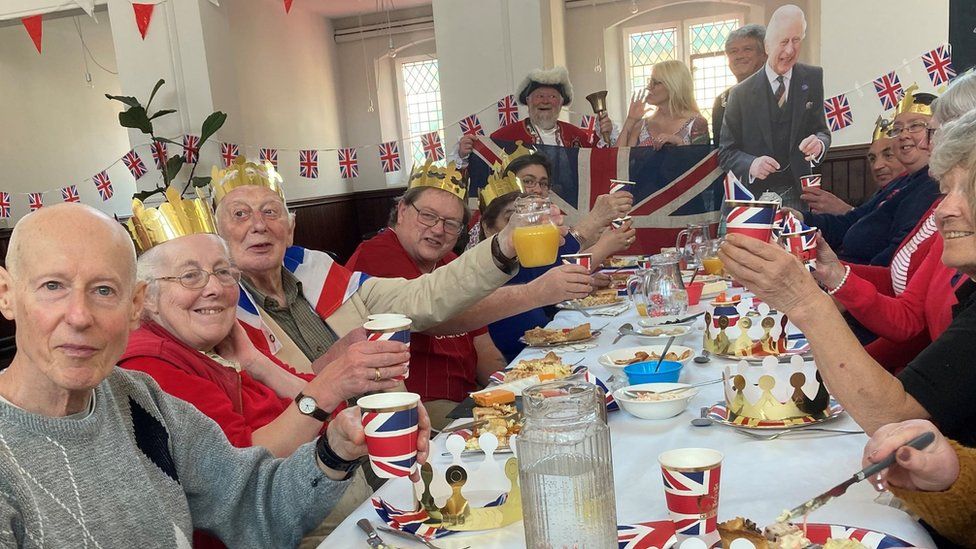 Long table with people raising a union jack cup as they tuck into a coronation lunch laid on by the Storehouse foodbank in Trowbridge