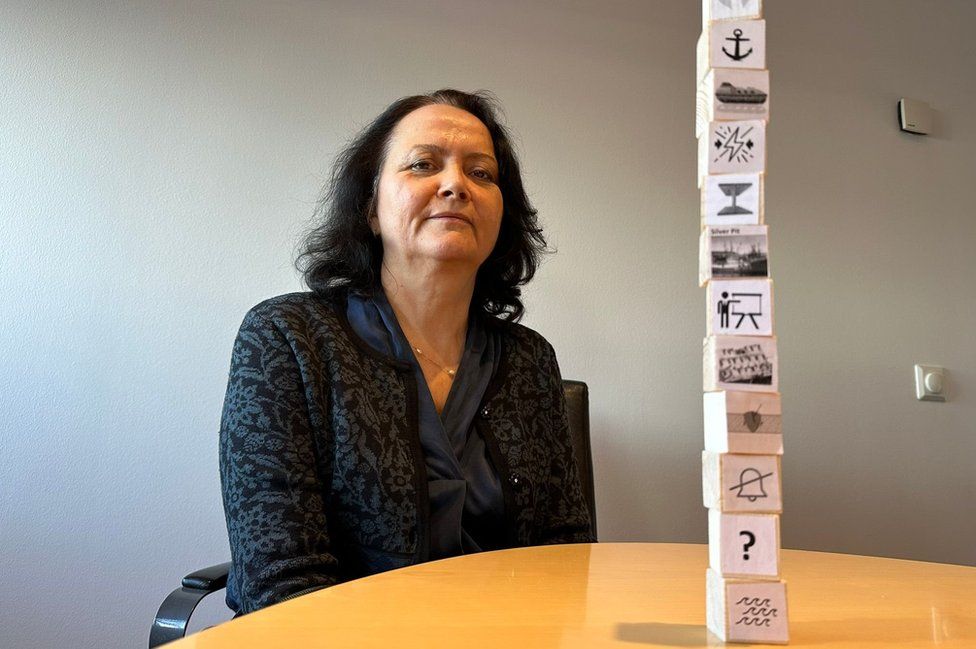 A woman sits next to a table on which are stacked 12 small wooden bricks each with a different image drawn on