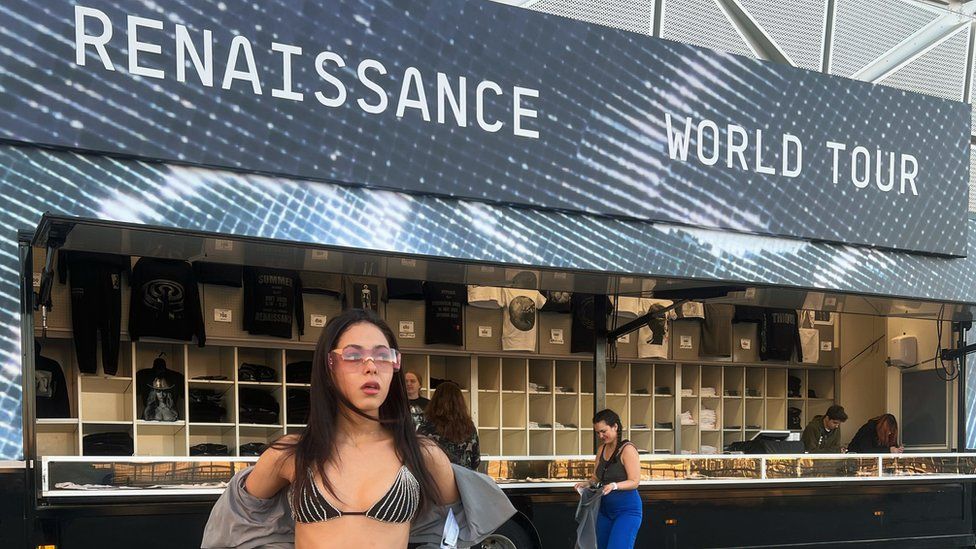 Ella is standing in front of the Renaissance World Tour merchandise stand