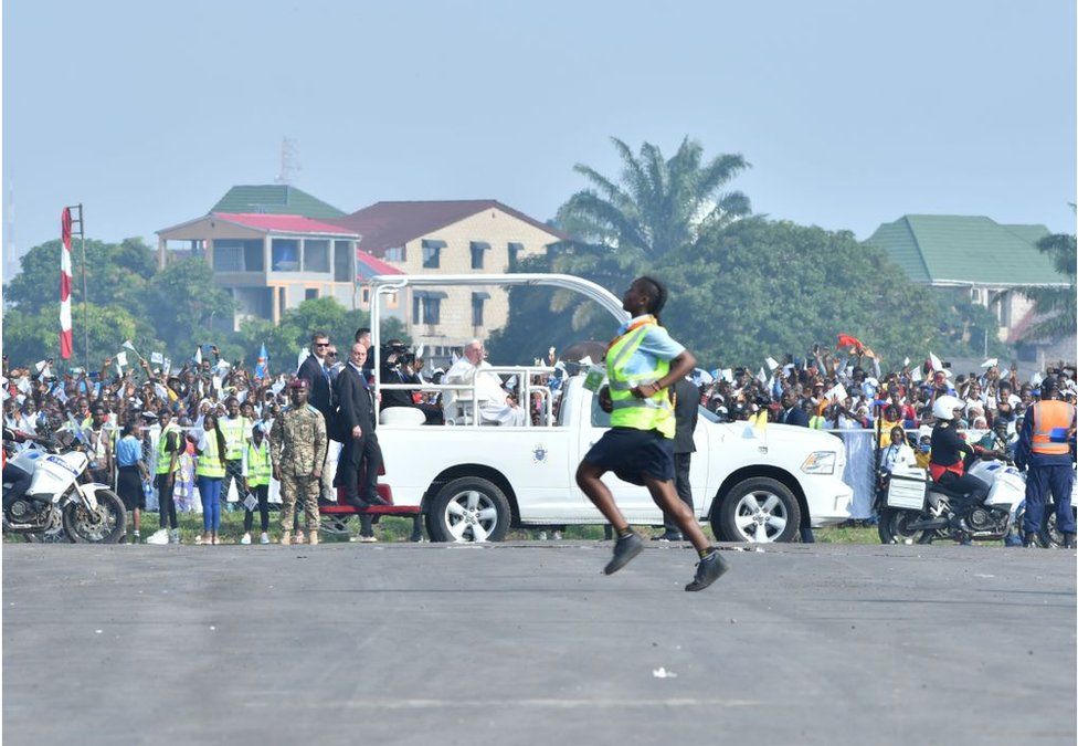 Woman, who looks like a steward, running in front of a crowd and the Pope Mobil.