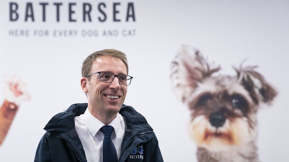 Battersea Cats and Dog Home's chief executive Peter Laurie smiles as he stands in front of a poster of a dog