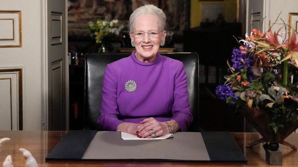 Queen Margrethe II, wearing a purple dress, smiles as she sits behind a desk