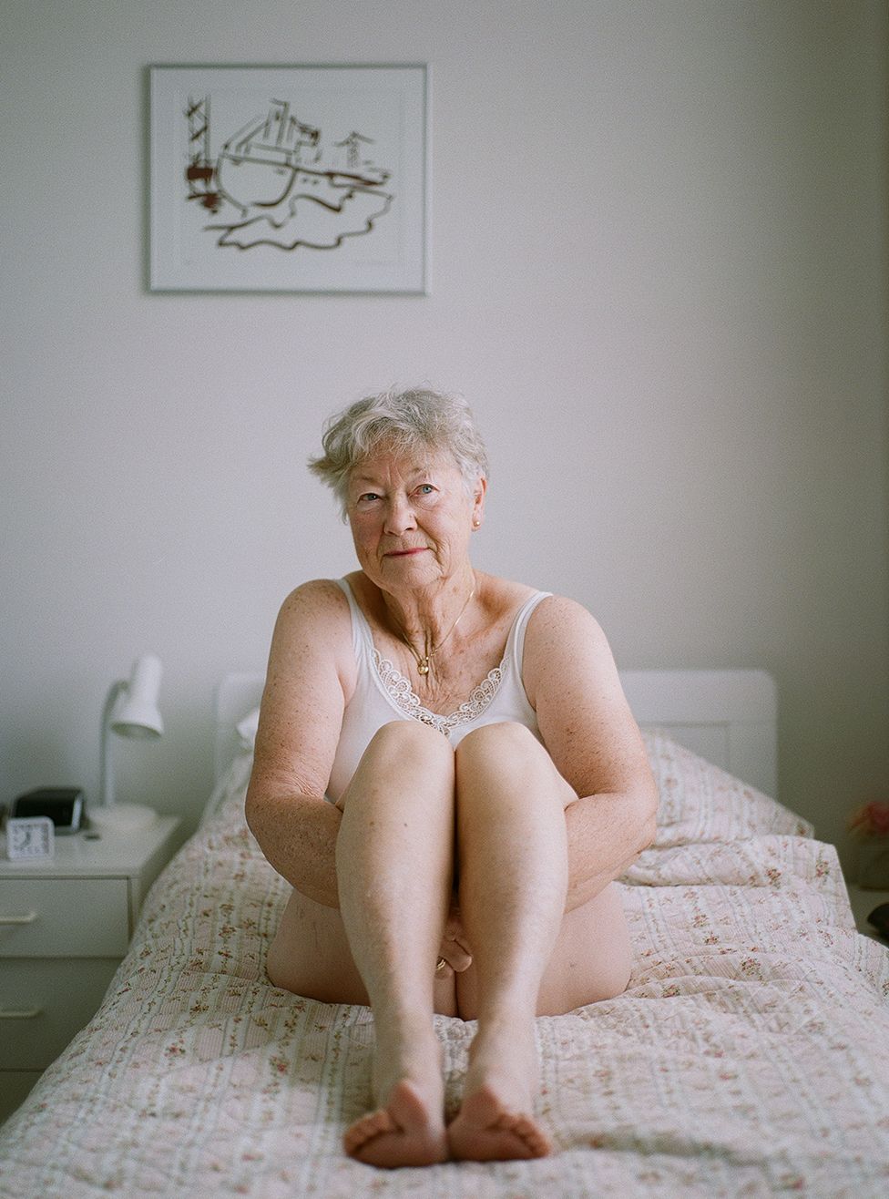 An elderly lady sits on a bed in her underwear