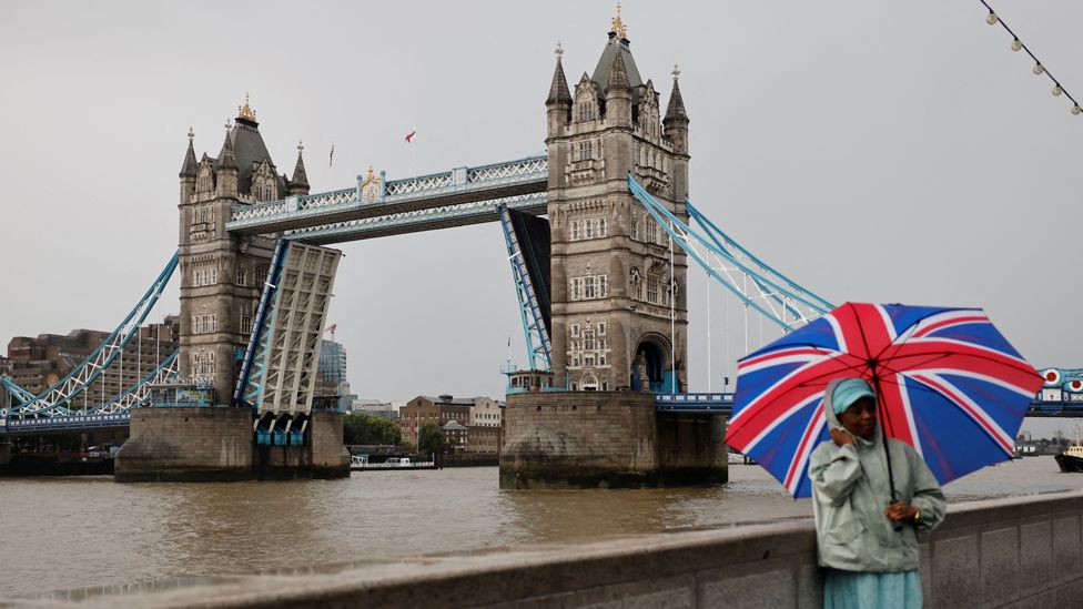 A man with a Union flag umbrella stands beside the stuck Tower Bridge