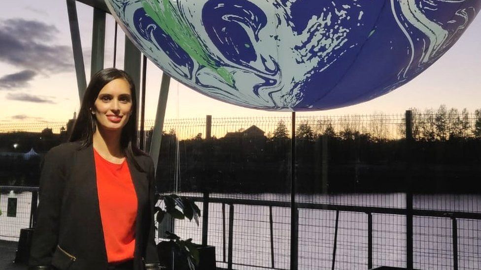 Amandeep standing infront of a blue, white and green big globe wearing a red top and a black blazer. It is sunset behind her with metal gates visible in front of some trees.