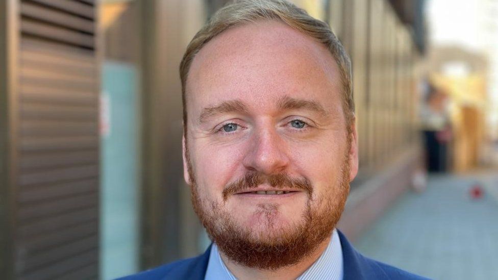 Man with short ginger hair and beard wearing suit and standing outside council offices