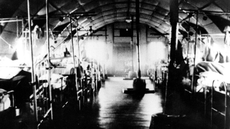 A slightly blurred black and white picture in which old hospital beds and stoves can be seen inside the hut, which has a domed roof.