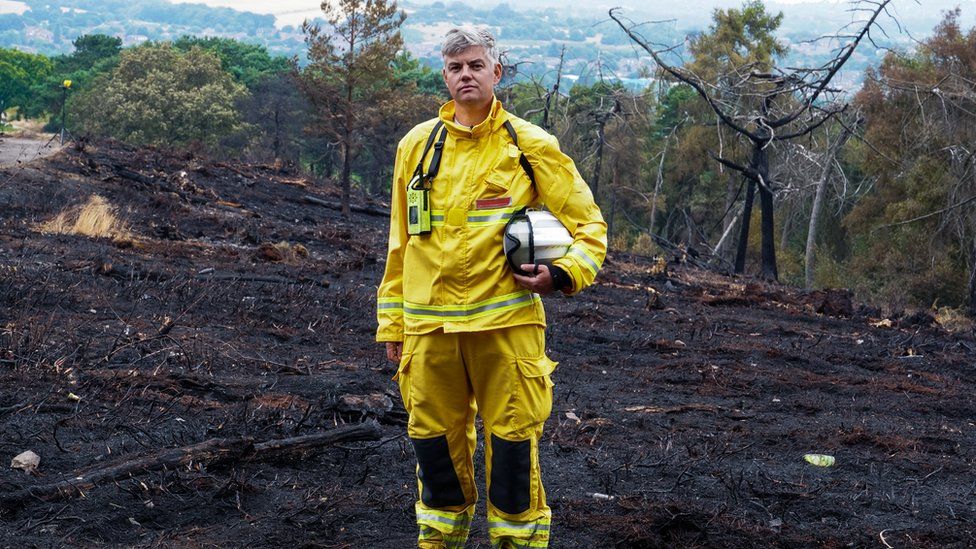 Dave Swallow standing in a yellow outfit against charred moorland.