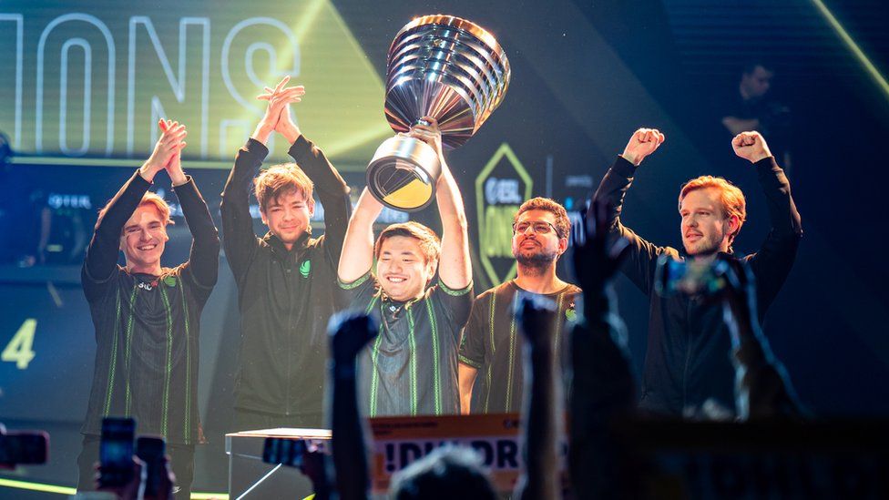 Five young men in matching long-sleeved tops stand in a line on-stage. In the middle, one of them holds an oversized, goblet-shaped trophy over his head with both hands. He's smiling widely as hands in the crowd before him go up in celebration. His team-mates have their arms raised, applauding or pumping the air with their fists, giving the shot a celebratory mood.