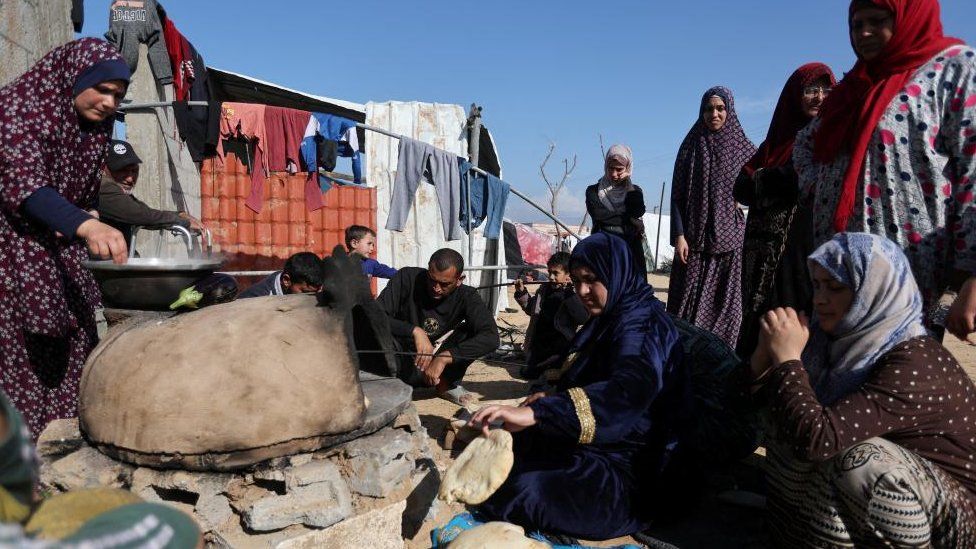 People wait while a woman prepares food, as displaced Palestinians, who fled their houses due to Israeli strike, shelter in a camp in Rafah, amid the ongoing conflict between Israel and Palestinian Islamist group Hamas, in the southern Gaza Strip, December 6, 2023.