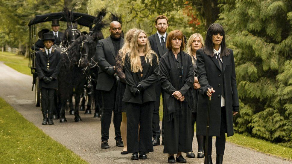 A still from episode 7 of The Traitors. Led by host Claudia Winkleman, the players walk as part of a funeral procession ahead of ushers and black horses pulling a carriage. All the players wear black and are pictured on a gravel path surrounded by greenery. Diane, a white woman in her 60s who has a short ginger bob, folds her hands as she stands behind Claudia. She is flanked by fellow players Molly, Evie. Paul and Miles.