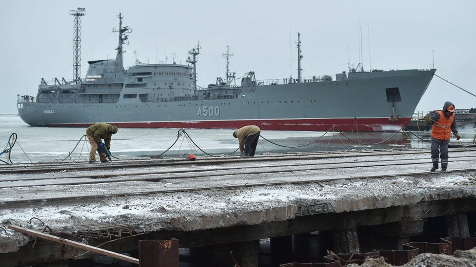 A very large military ship is seen floating on an icy bay in the background, while closer to the camera, a handful of men work on a long, empty pier area