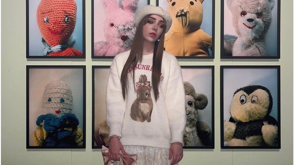 Kellen is standing in front of eight framed pictures of different animal teddy bears. She is wearing a white knitted jumper that features a picture of a brown rabbit with a pink bow tied around its ears. She has long brown straight hair and is wearing a white beret with a small group of pearls on the left hand side of it. Kellen is posing side on and is looking down towards the floor rather than towards the camera.
