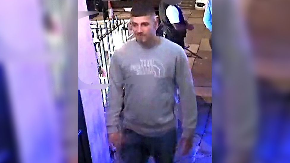 CCTV image of a man in a bar. He has dark hair and is wearing a grey North Face jumper with dark blue jean