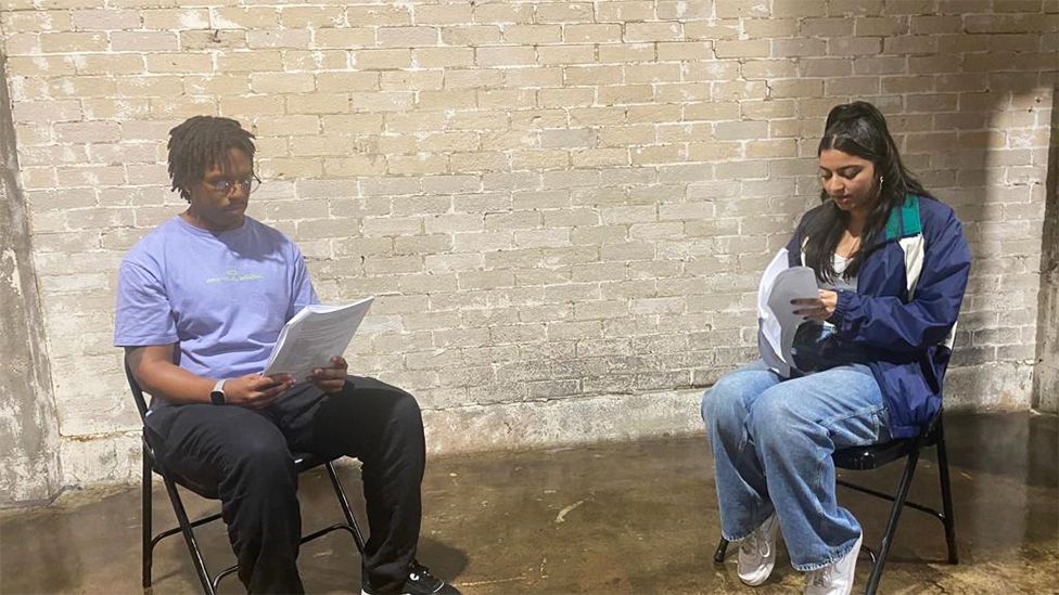 Siyani rehearsing with another member of the cast. They are both sat down on black metal chairs. Siyani is on the right wearing a navy blue jacket, white top and light blue jeans, facing the other cast member who is on the right wearing a purple top, dark jeans, watch on his right wrist, black shoes and glasses. The background is brick wall, discolourd with white marks.