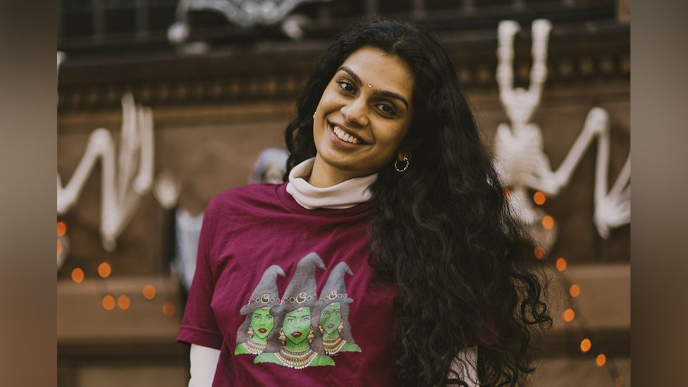 A young South Asian woman sits in front of a brownstone building that's decorated with white halloween skeletons, orange pumpkin lights and other spooky decorations. She's wearing a burgundy t-shirt with a design showing three witches. They have the traditional green skin and pointy black hats associated with witches, but they wear South Asian-style necklaces, ornate gold earrings and they have jewellery on their foreheads. Their hats are also decorated with a pattern of multicoloured hoops. The young woman wearing the t-shirt is smiling, and herself wears gold hoop earrings and sports a jeweled bindi on her forehead. Her long, dark hair cascades down past her shoulders.