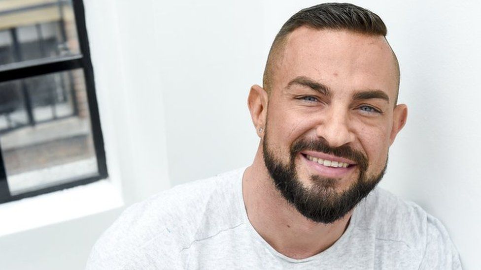 Robin Windsor poses against a white wall, there is a corner of a window in the top left of the photo. He is smiling and wearing a white long sleeved t-shirt.