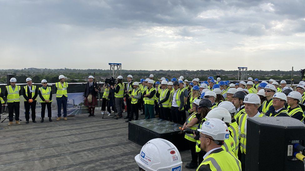 Scores of people wearing hi-viz vests and white hard hats at a topping-out ceremony
